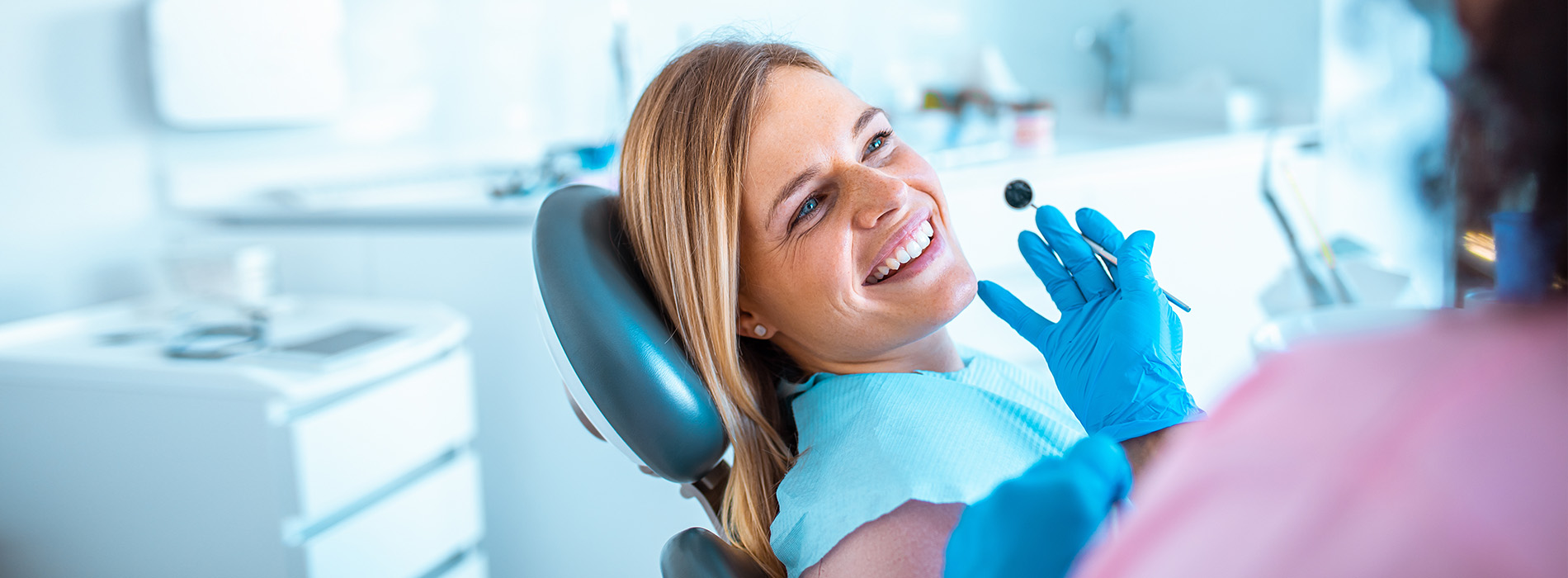 A woman seated in a dental chair, smiling and looking towards the camera while a dental professional attends to her.