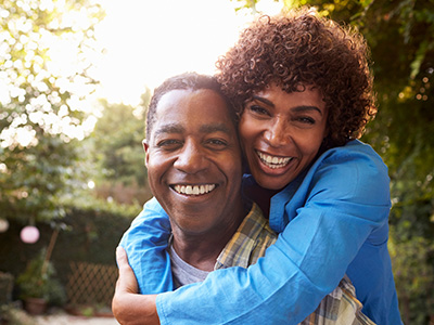 A man and woman, both smiling, embrace each other in a warm hug with a garden backdrop.