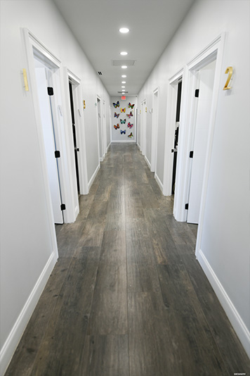 Long, narrow hallway inside a building with light-colored walls and floor, featuring a series of closed doors on the left side.