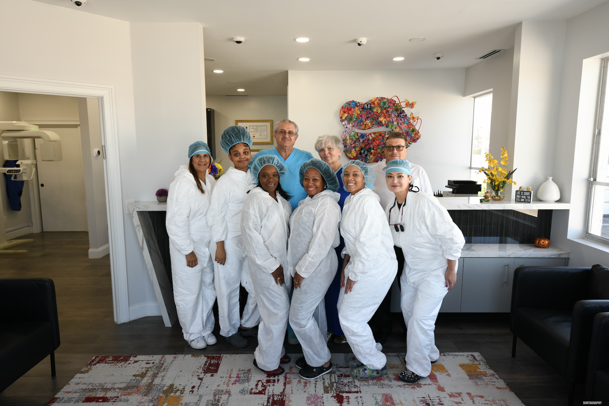 The image shows a group of individuals, likely healthcare professionals given their attire and the setting that appears to be a medical facility, standing together in front of a desk with a laptop.