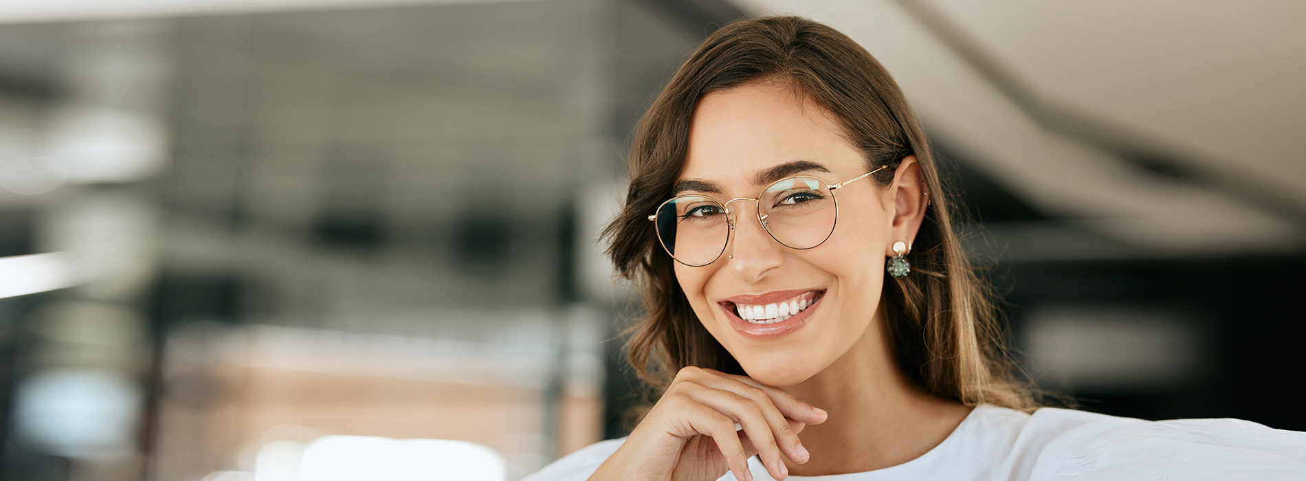The image is a photograph of a smiling woman with glasses, wearing a white top and her hair styled down. She appears to be in an indoor setting with reflective surfaces behind her.