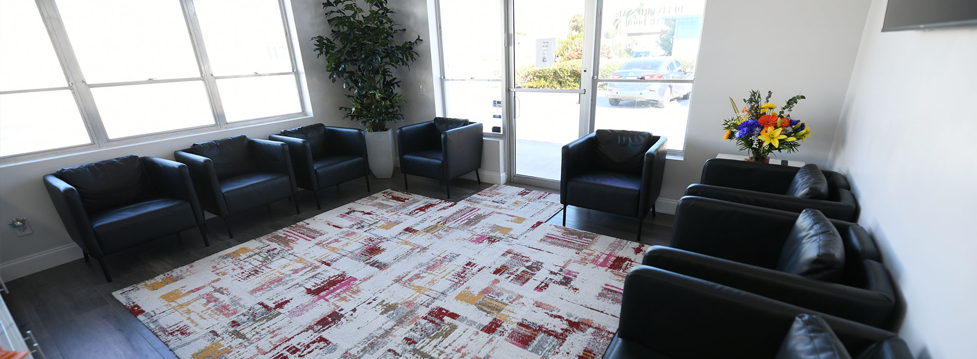The image shows an interior space that appears to be a waiting or reception area, furnished with multiple black leather chairs arranged around a central area rug with a pattern of intersecting lines in shades of red and white.