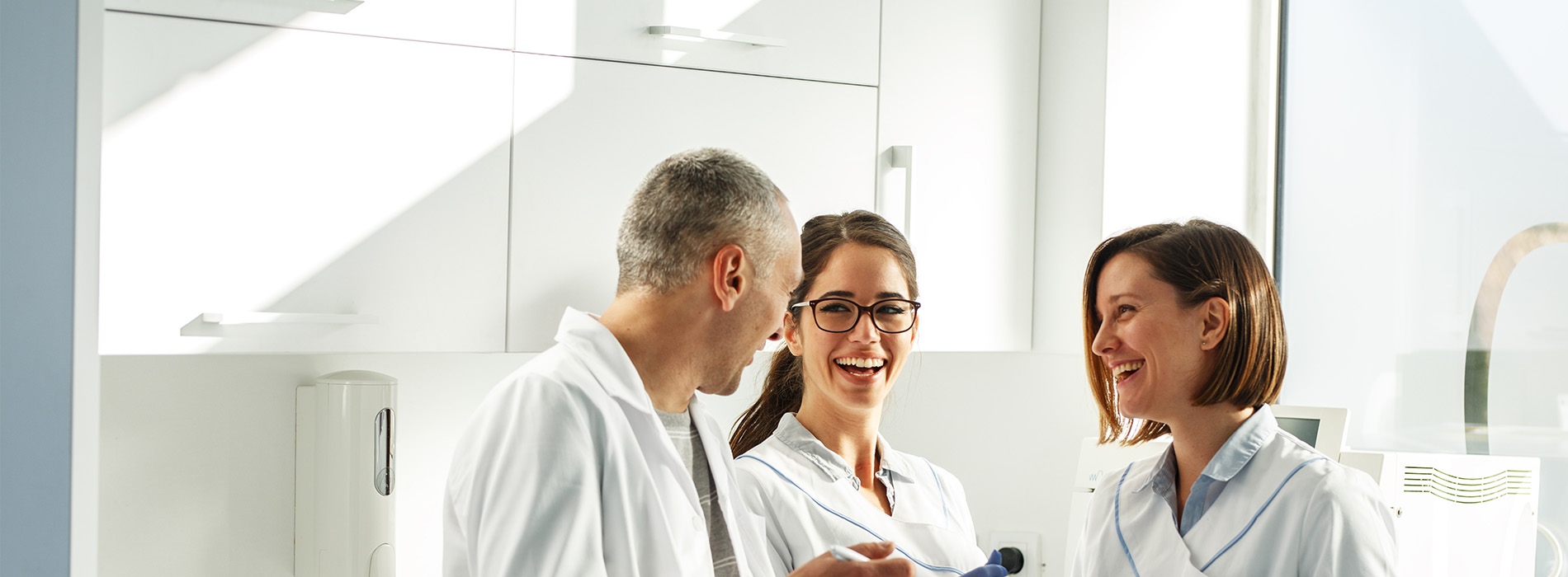 Three individuals in a professional setting, possibly a dental or medical office, with one person smiling at the camera and another holding a tablet, all wearing white lab coats.