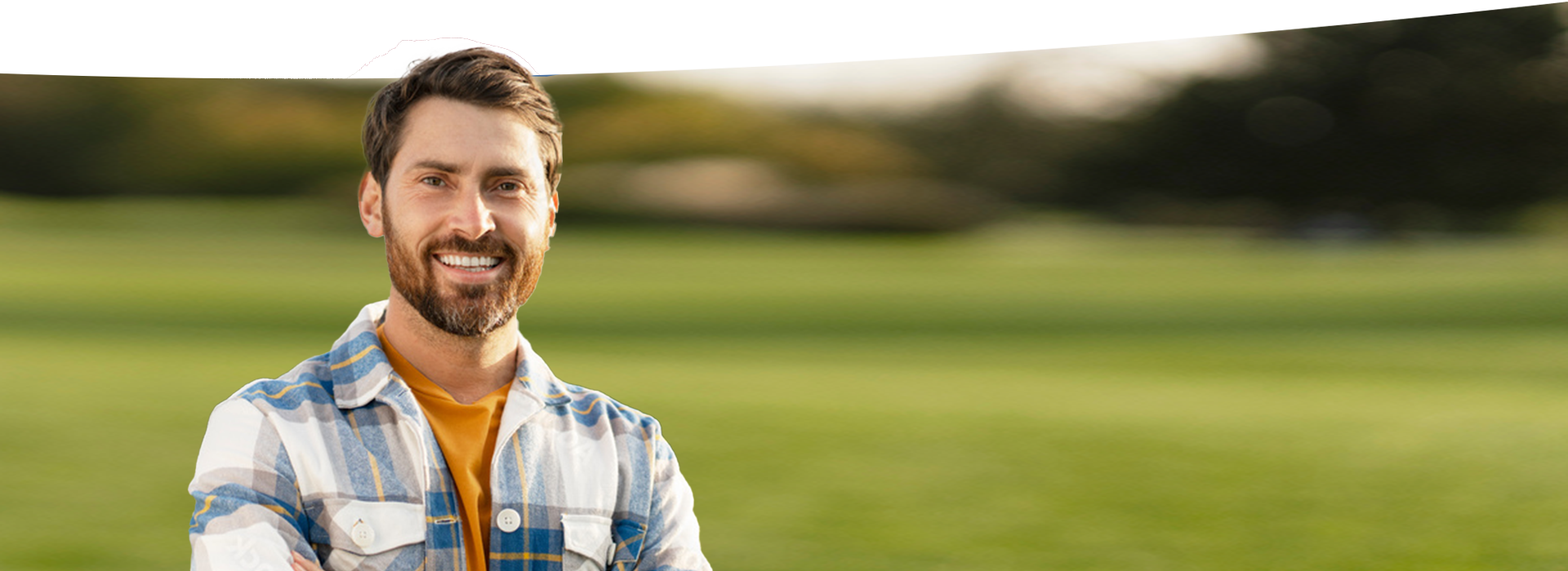The image shows a man standing on a grassy field, smiling at the camera. He is wearing a plaid shirt and has short hair.