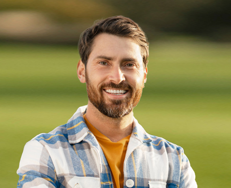 The image features a man with facial hair, smiling at the camera. He is wearing a plaid shirt and appears to be standing outdoors on a golf course.