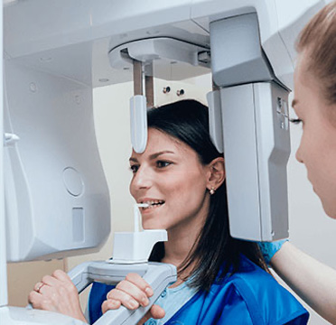 Woman in blue scrubs under a large, white 3D scanner, with another woman assisting.