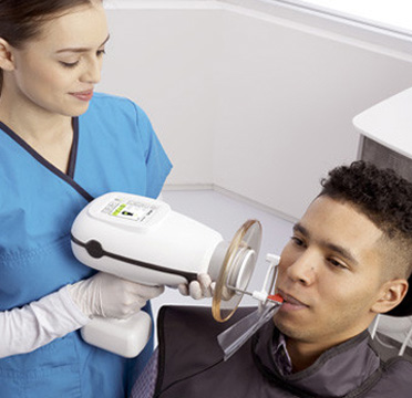 A man in a dental chair receiving oral care from a professional using an electronic device.