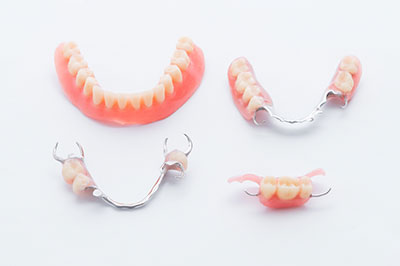 The image displays a set of dentures with a full upper and lower arch, along with a pair of partial upper dentures, all on a plain white background.