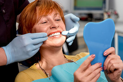 The image shows a woman sitting in a dental chair, smiling at the camera, with a dental hygienist adjusting her mouthpiece. She is holding a blue dental impression tray while wearing gloves and a face mask.
