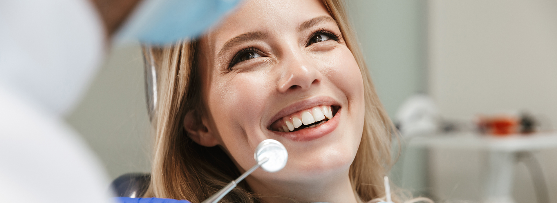 The image features a woman with a bright smile, looking at the camera, while seated in a dental chair during a dental examination.