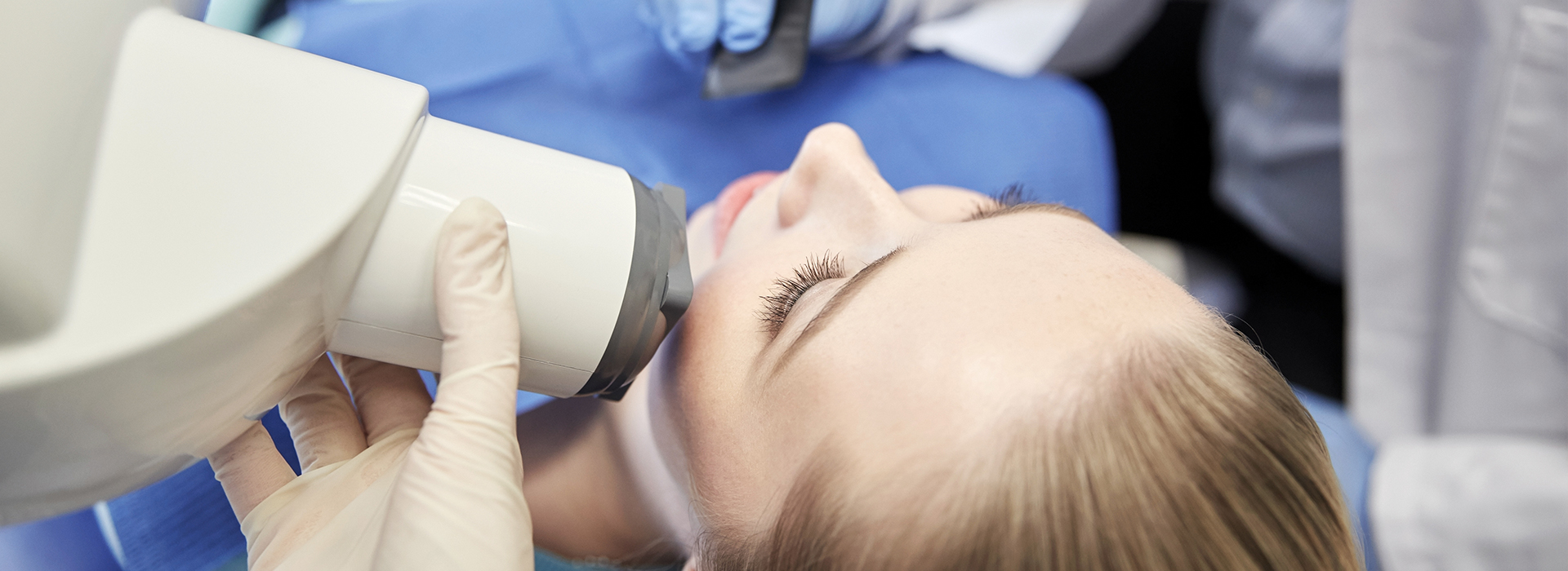 The image shows a person receiving dental care, possibly an examination or cleaning, while seated in a dental chair with a dental professional's hands visible near their face.