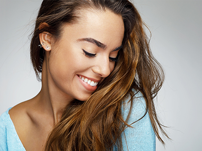 The image shows a woman with long hair, smiling and looking downwards. She is wearing a light blue top and has her hair styled in loose waves.
