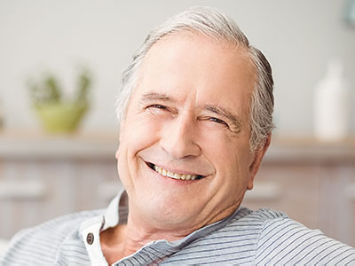 A smiling older man with gray hair, wearing a blue shirt, seated in a living room.